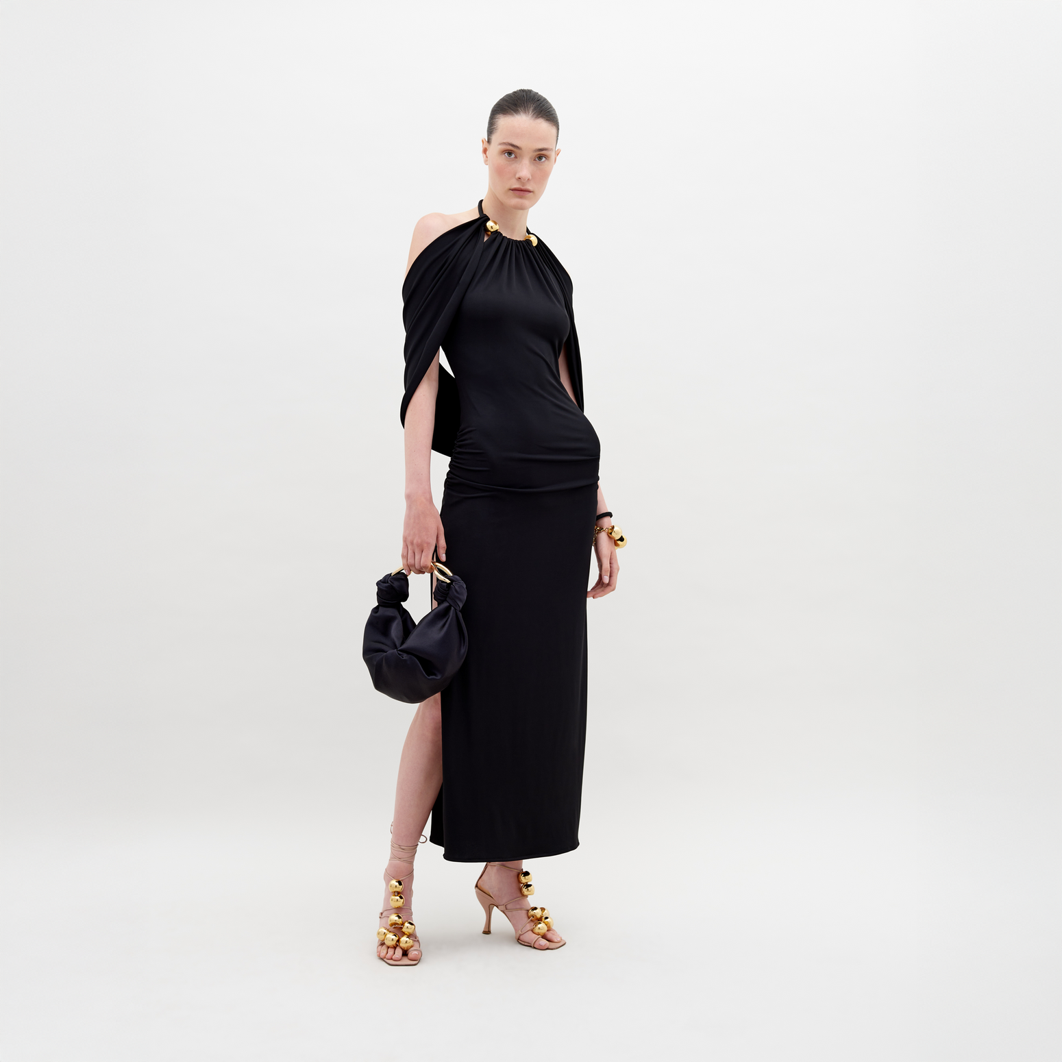 A woman stands against a white background, showcasing the Odele Dress Black with gold accents. She holds a small black bag and wears gold-embellished high heels. Pre-order now to ensure delivery by November 15th, 2024.