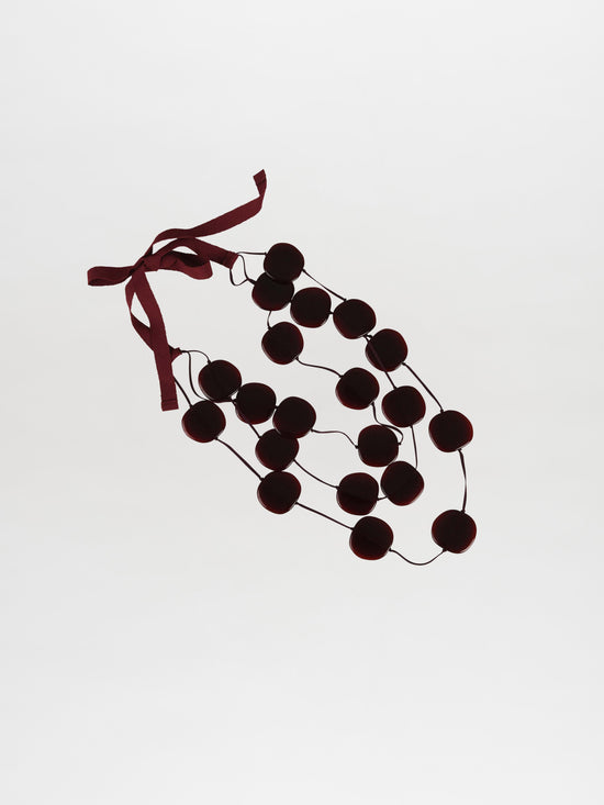 The Olivia Necklace in Burgundy showcases a stunning acrylic statement design, featuring dark round beads crafted from exquisite Italian resin. These are elegantly strung together with a maroon ribbon, and the rich burgundy hue of the beads stands out beautifully against the white background.