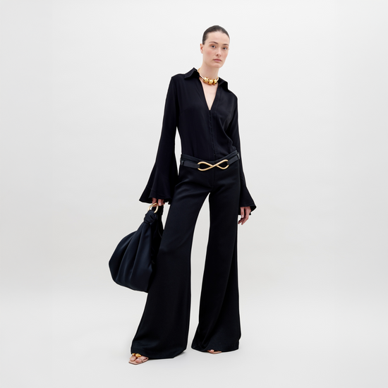 A person stands against a plain background wearing a stylish, sleeveless black top with draped fabric and Ovidia Pant Black, accessorized with gold-toned bracelets and sandals. Available for pre-order now, this ensemble will ship by date in early 2024.