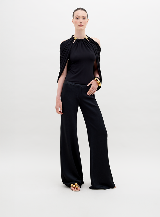 A person stands against a plain background wearing a stylish, sleeveless black top with draped fabric and Ovidia Pant Black, accessorized with gold-toned bracelets and sandals. Available for pre-order now, this ensemble will ship by date in early 2024.