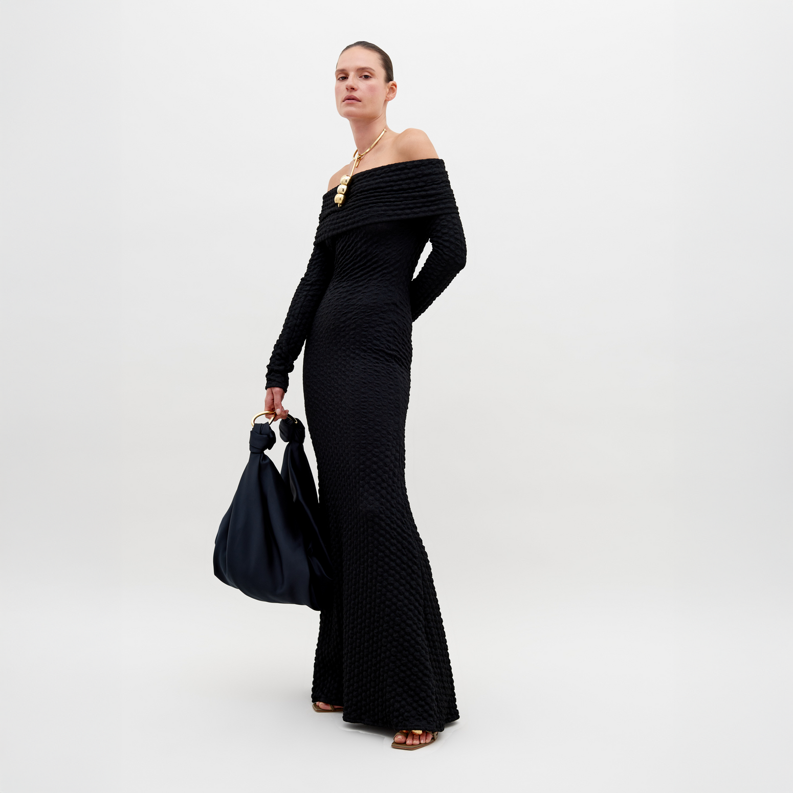 A person stands wearing a long, off-the-shoulder black textured Ozara Dress Black, holding a large black bag. They have a gold necklace and their hair is pulled back. The background is plain white, perfect for promoting the new collection available for pre-order. Mark your calendars for the ship by date: November 15th, 2024.