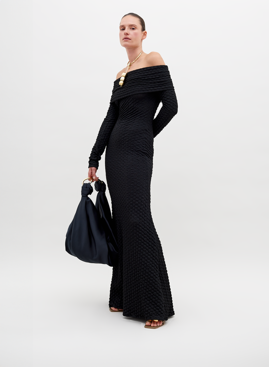 A person stands wearing a long, off-the-shoulder black textured Ozara Dress Black, holding a large black bag. They have a gold necklace and their hair is pulled back. The background is plain white, perfect for promoting the new collection available for pre-order. Mark your calendars for the ship by date: November 15th, 2024.