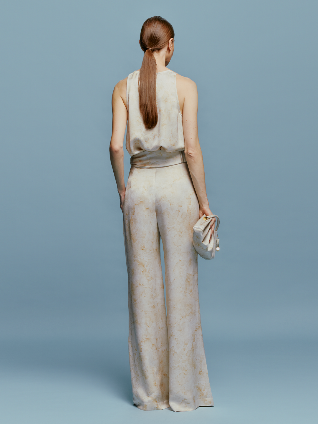 A woman stands wearing the Palermo Pant in Cream Abstract Print, a strapless jumpsuit featuring a light-colored fabric and a floral design on the top, accessorized with a small clutch against a plain blue background. Pre-order now for August shipping dates and elevate your summer wardrobe effortlessly.