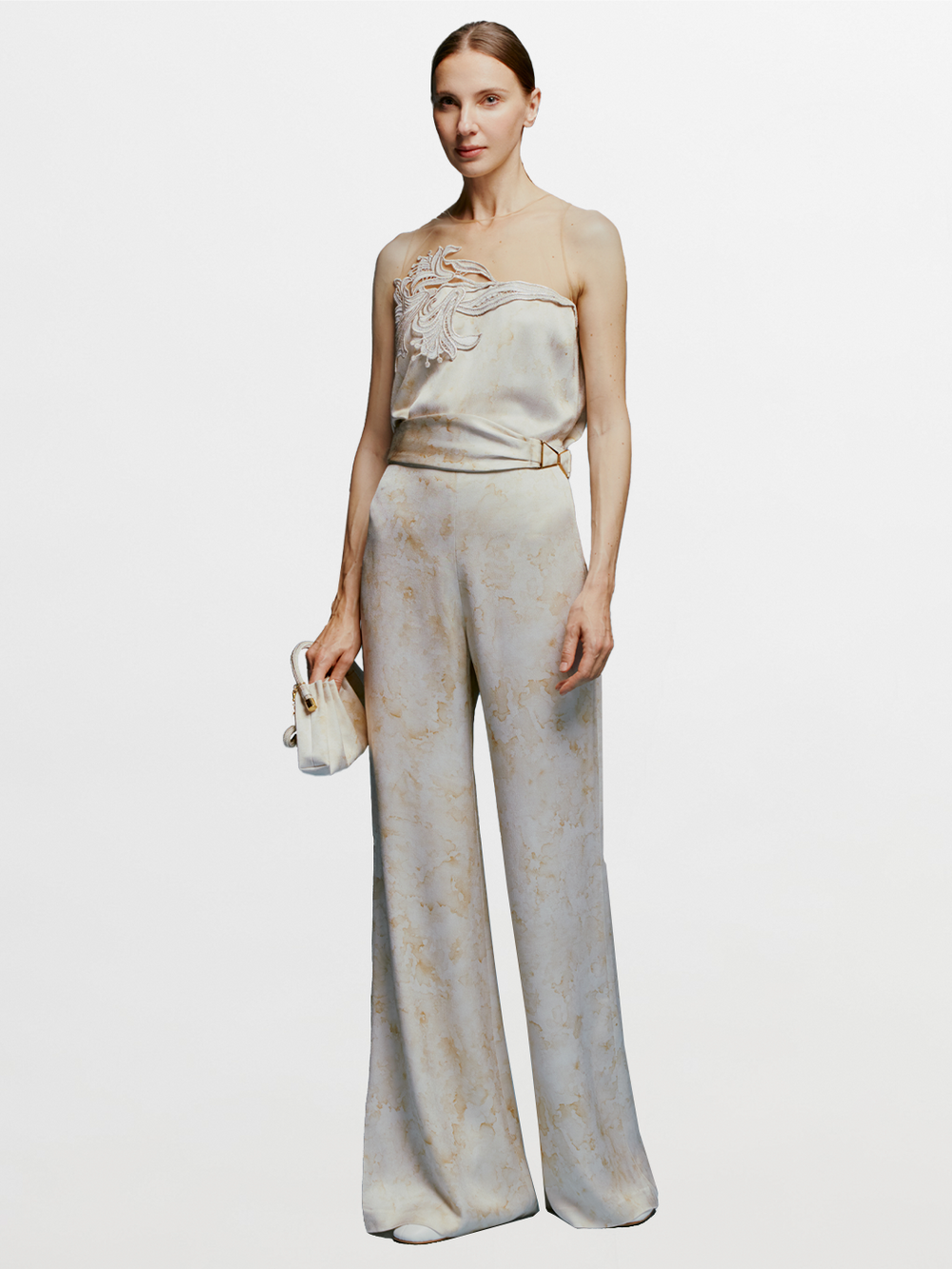 A woman stands wearing the Palermo Pant in Cream Abstract Print, a strapless jumpsuit featuring a light-colored fabric and a floral design on the top, accessorized with a small clutch against a plain blue background. Pre-order now for August shipping dates and elevate your summer wardrobe effortlessly.