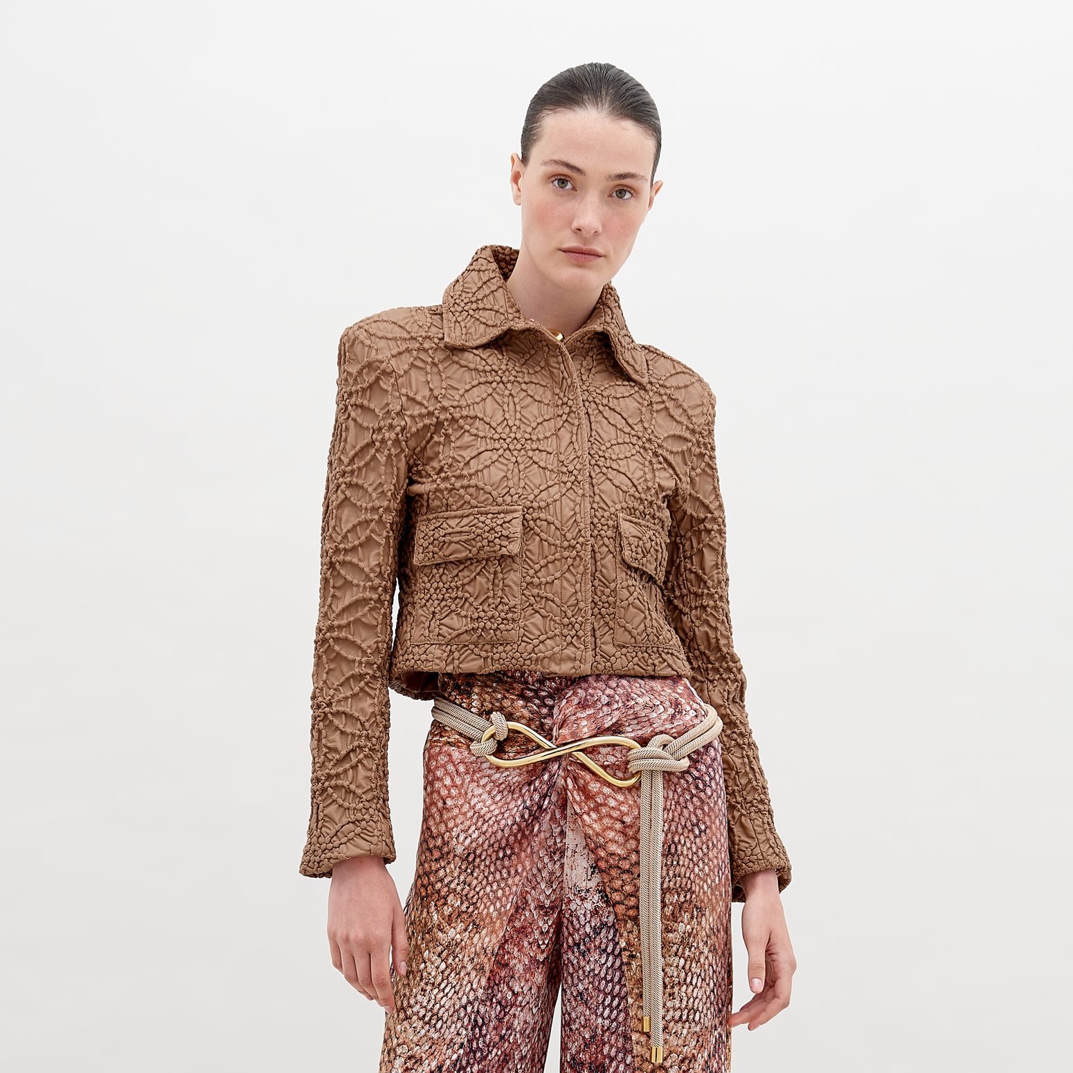 A woman stands against a plain background, wearing the Perla Jacket Brown and patterned high-waist pants. A decorative knot belt accentuates her waist. This stylish ensemble is available now, ensuring you can enhance your wardrobe immediately.