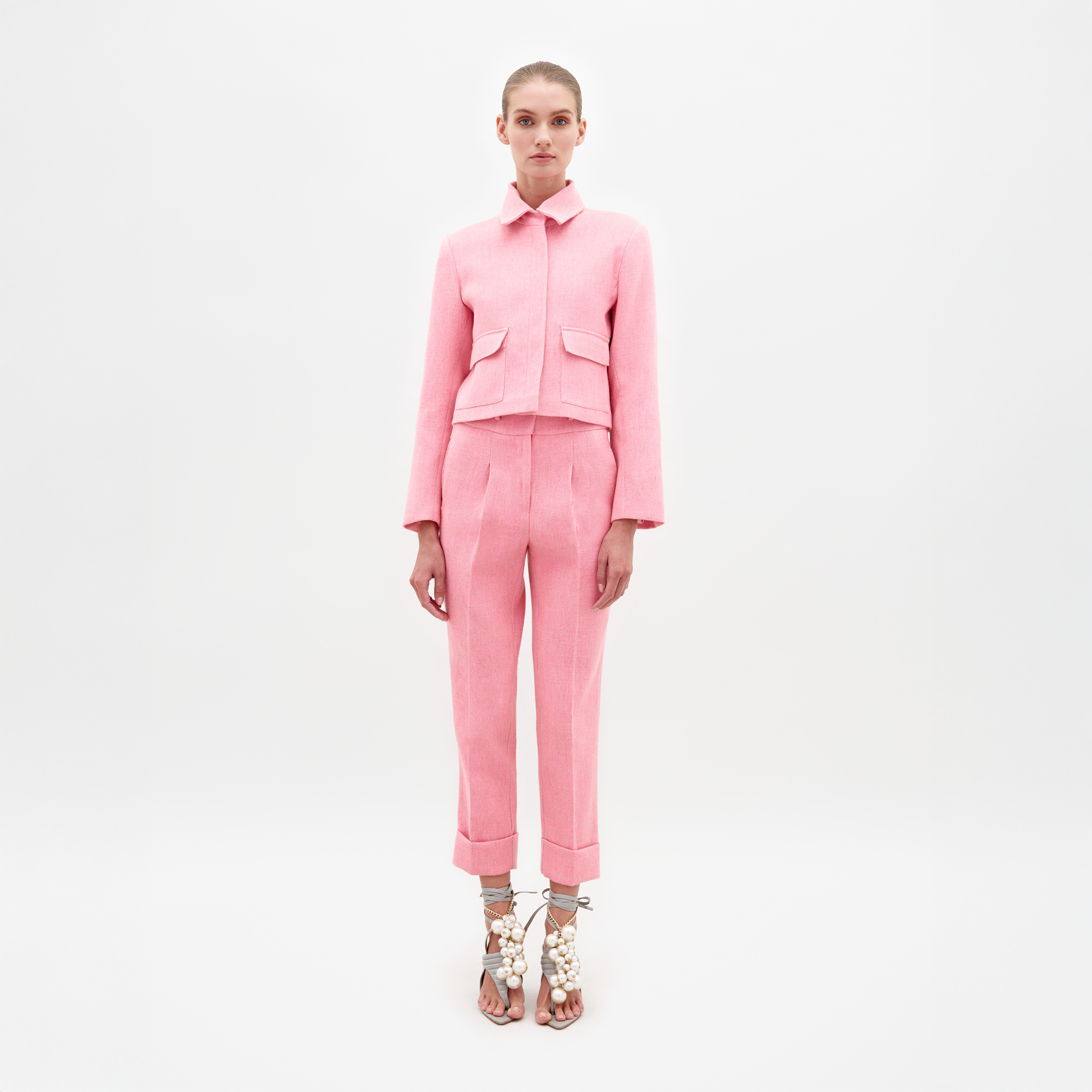 A person stands against a plain background, wearing the Moad Pant Pink suit, featuring a tailored cropped jacket and matching pink pants. They complete their look with high-heeled shoes adorned with white embellishments, ready for shipment by February 15th, 2025.