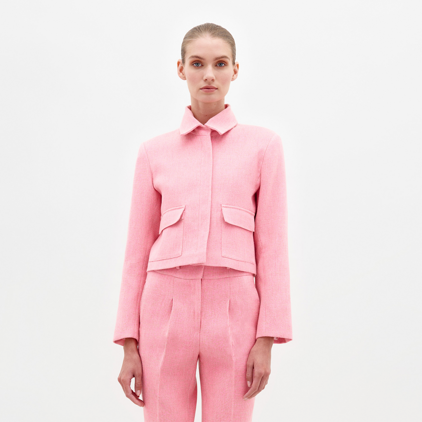 A person with tied-back hair is wearing a matching pink tailored suit, including the Perla Jacket Pink, which features a collar and two front pockets, standing against a plain white background. This stylish look will be available for pre-order, with the ship date set for February 2025.