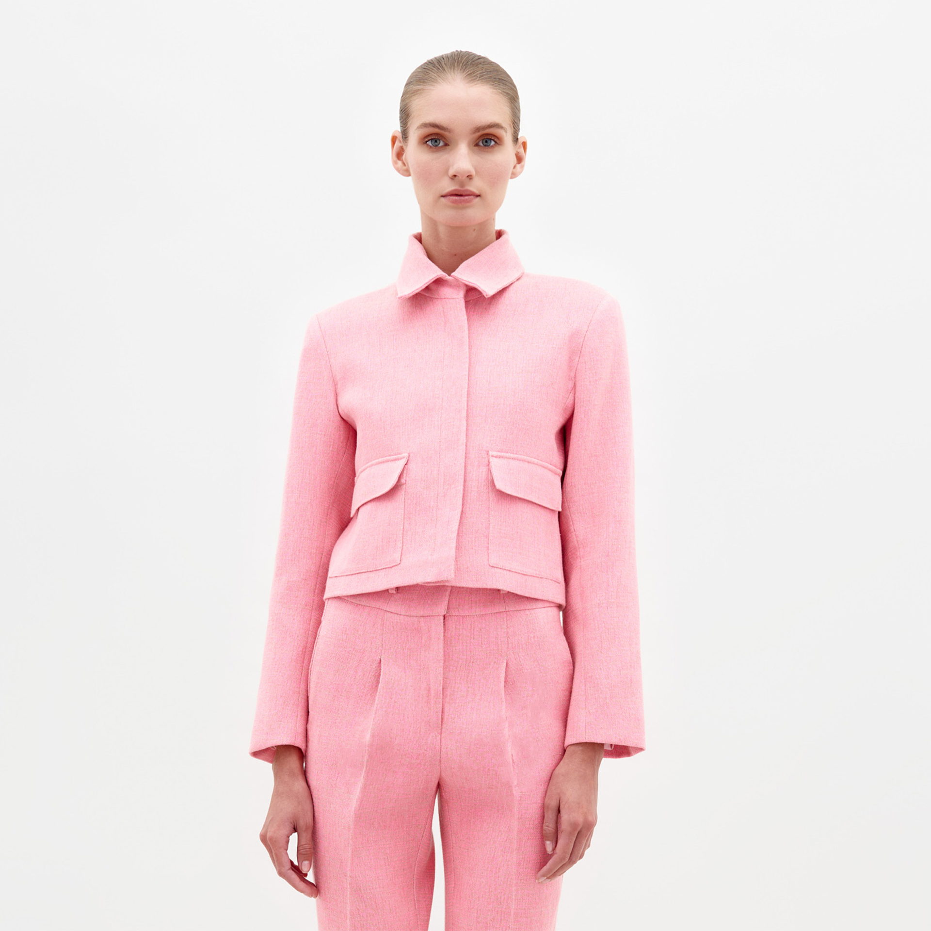 A person with tied-back hair is wearing a matching pink tailored suit, including the Perla Jacket Pink, which features a collar and two front pockets, standing against a plain white background. This stylish look will be available for pre-order, with the ship date set for February 2025.