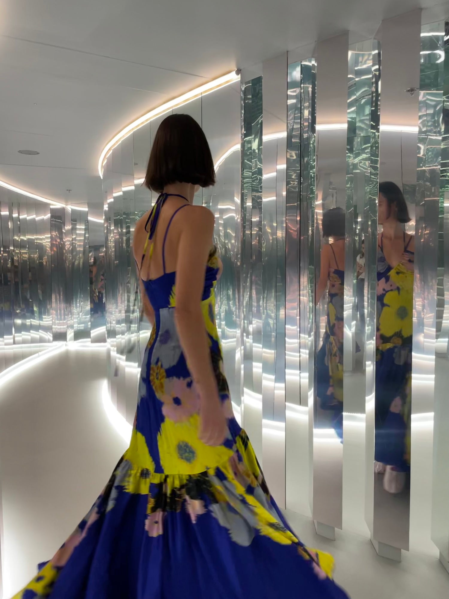 Person in a blue dress with yellow flowers walks through a hallway lined with mirrored walls.