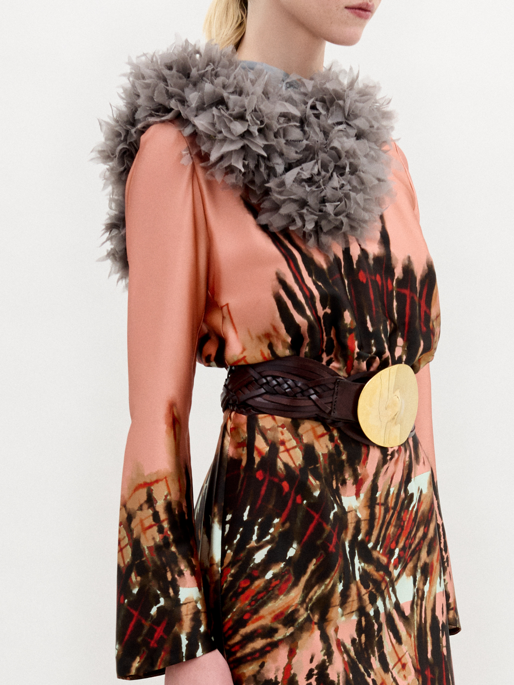 A person stands in profile wearing the Pesaro Dress—a peach silk midi with a large feathered design, wide belt with round buckle, gray fur collar, and black heeled boots—set against a plain background.