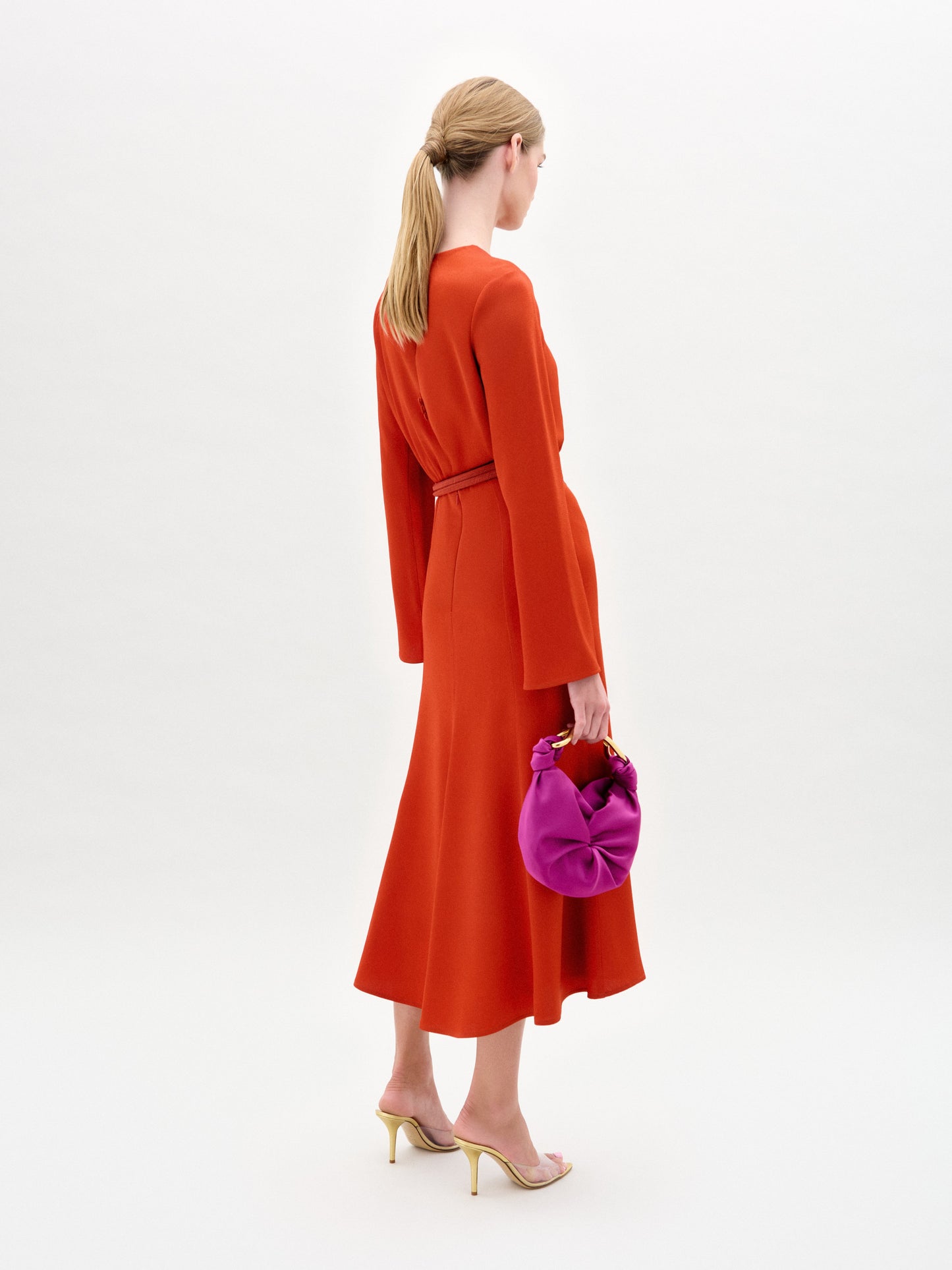 Sporting the Pesaro Dress, a long-sleeved orange midi crafted from luxurious Italian fabric with an abstract print, the woman holds a small purple bag. Her hair is styled back, and she finishes her look with chic open-toed heels.