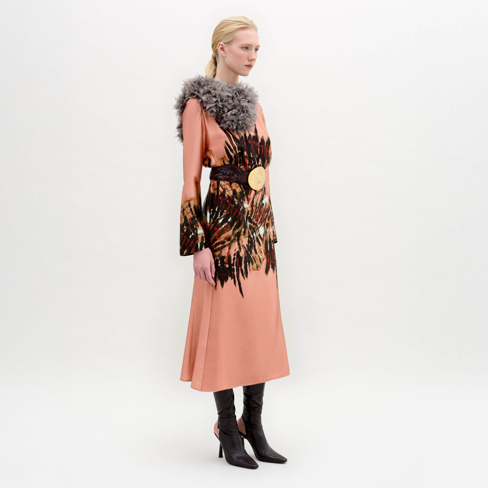 A person stands in profile wearing the Pesaro Dress—a peach silk midi with a large feathered design, wide belt with round buckle, gray fur collar, and black heeled boots—set against a plain background.