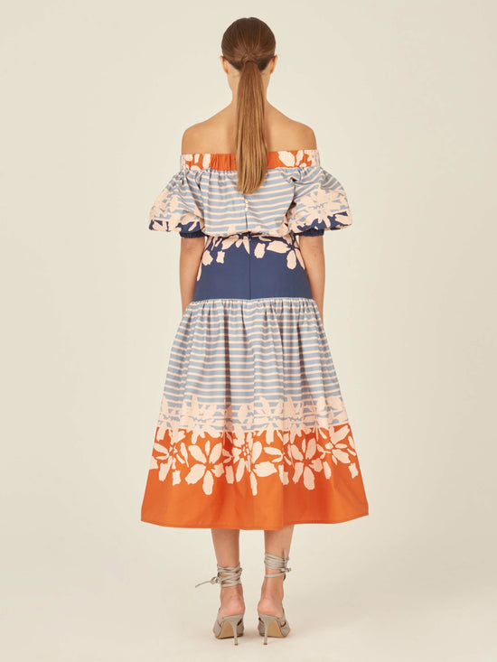 The Fulvia Dress Celeste Orange captivates with an off-the-shoulder silhouette, accentuated by blue stripes and a navy waistband. The dress features charming floral patterns in peach and orange on the top and bottom sections, striking a delightful balance between elegance and playful style.