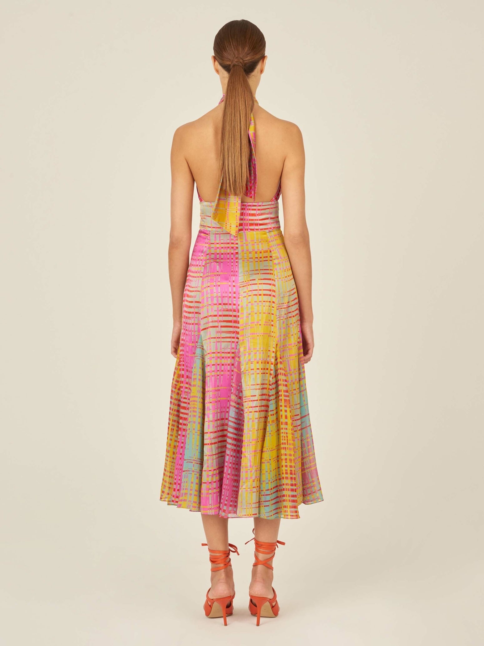 A person wearing a vibrant Silvana Dress White Digital with a twist-halter silhouette, showcasing a plaid pattern and paired with orange strappy sandals.