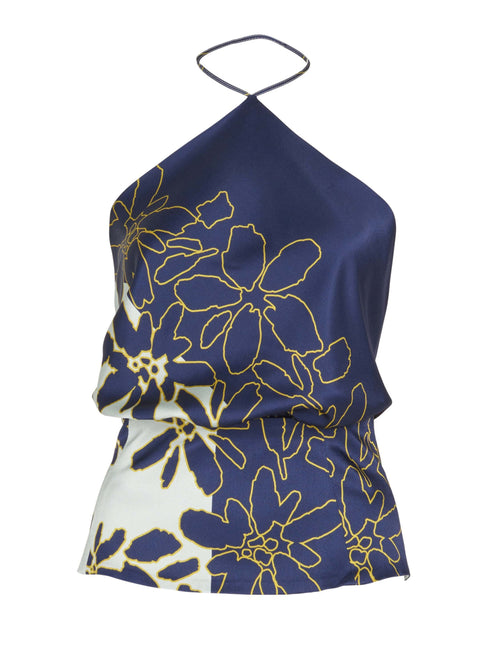 A person with long hair stands facing away, showcasing the Chantal Blouse Navy Citrine, an open back halter top featuring blue and white floral silk patterns, paired stylishly with yellow wide-leg pants.