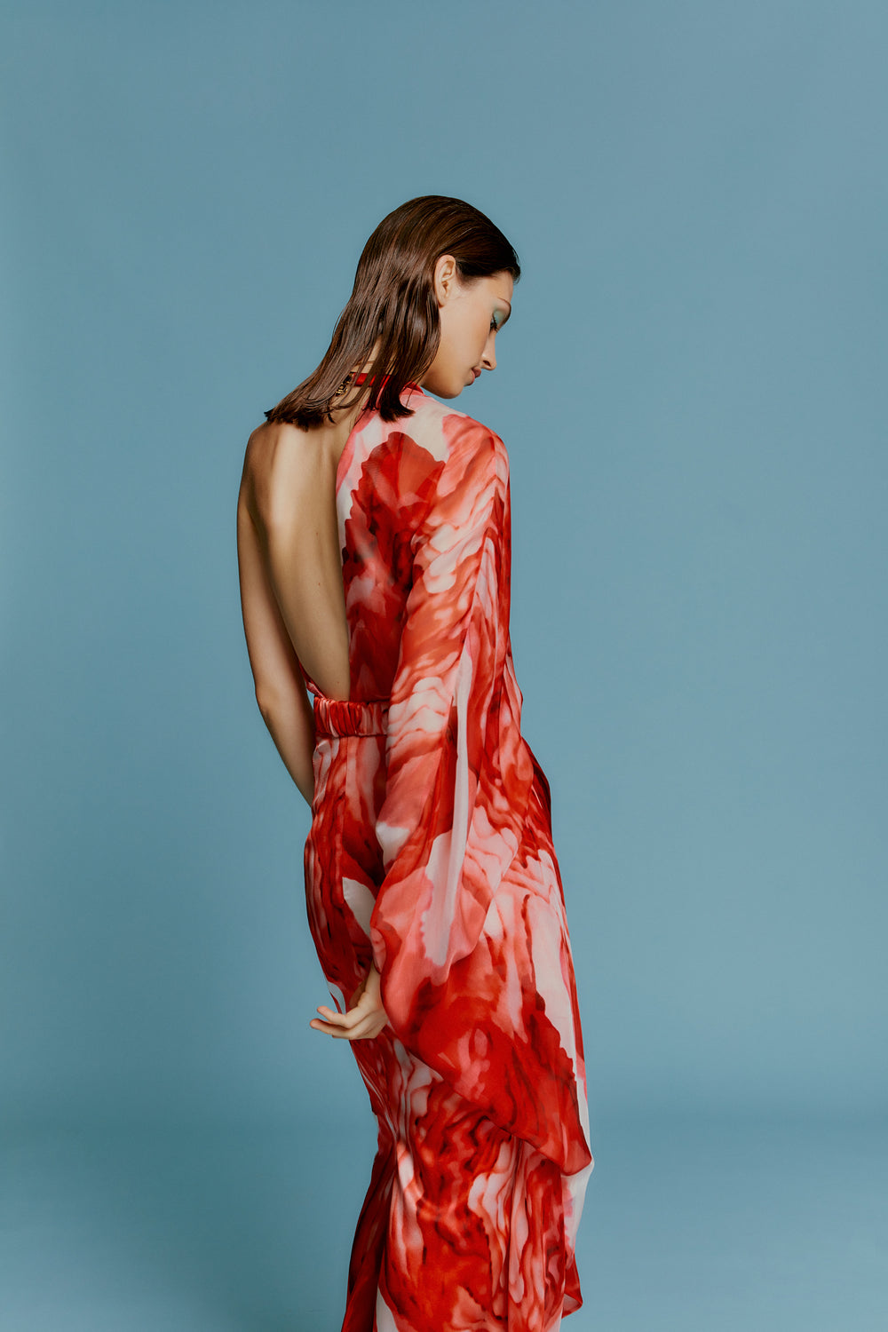 A woman stands with her back turned, wearing a red and white patterned, backless dress against a blue background.