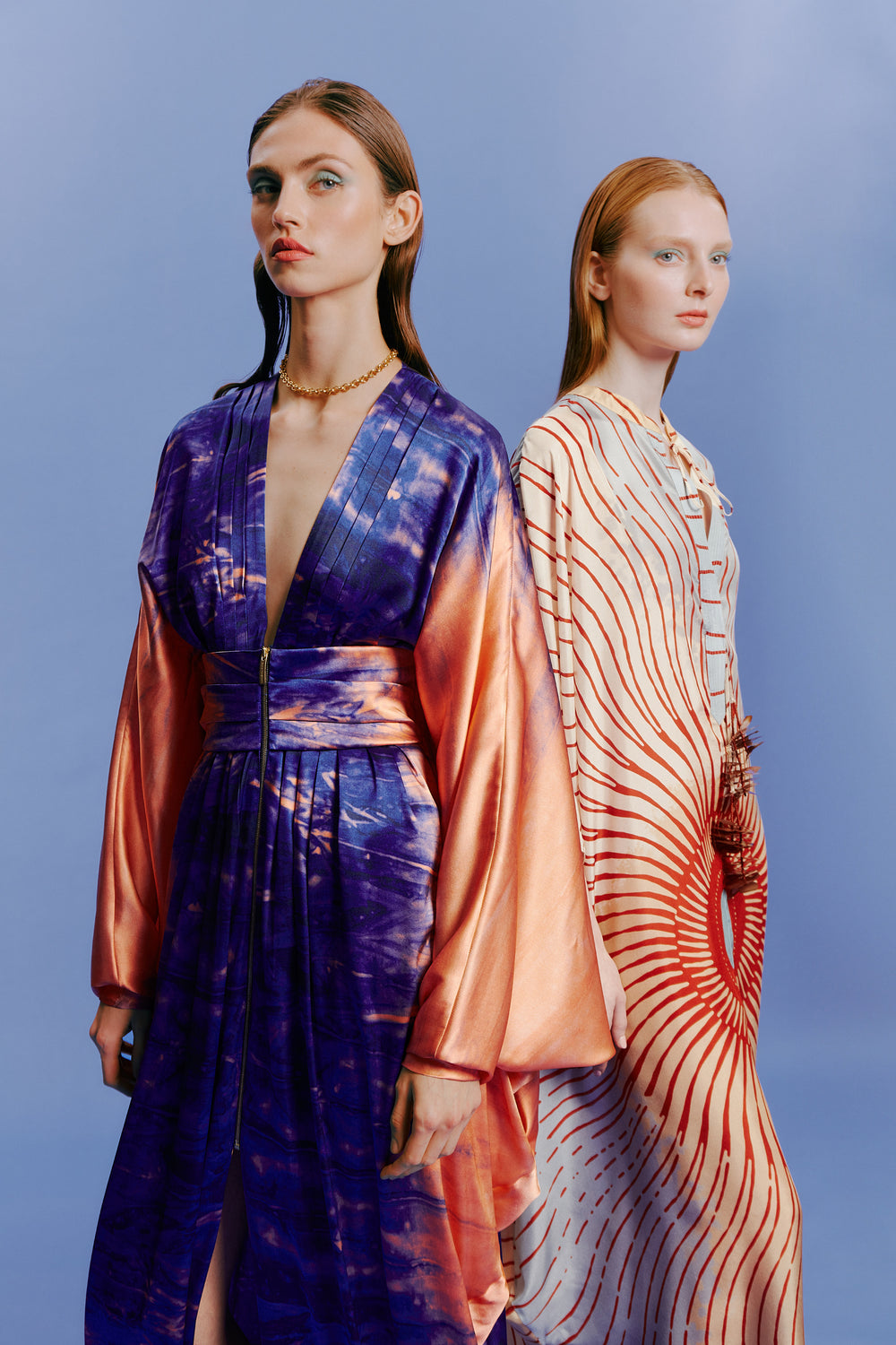 Two models in colorful, flowing outfits pose against a blue background. The first wears a purple and orange dress, while the second model is in a patterned red and white dress.
