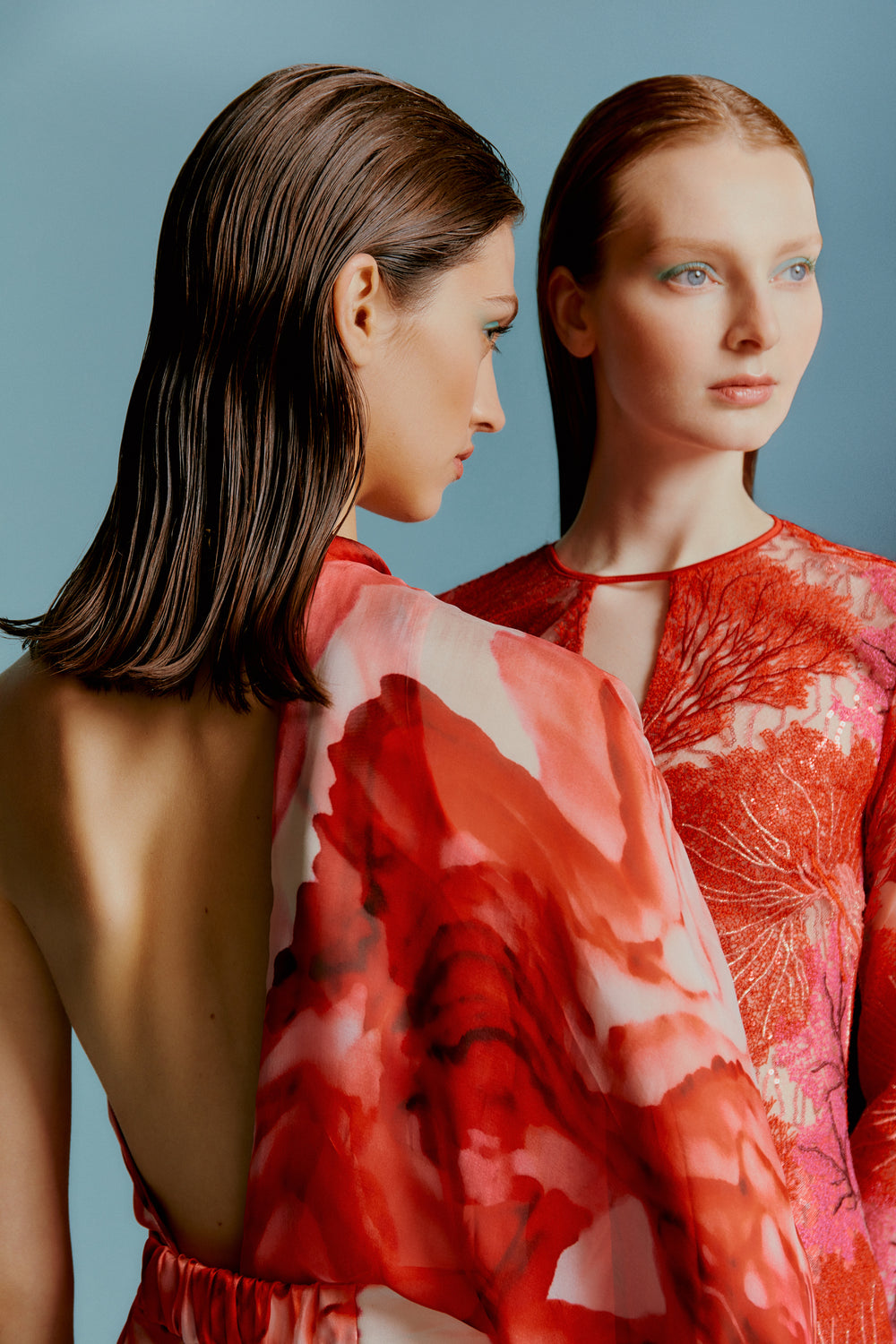 Two women with wet-look hairstyles wear elegant red floral dresses against a soft blue background. One faces forward; the other is in profile.