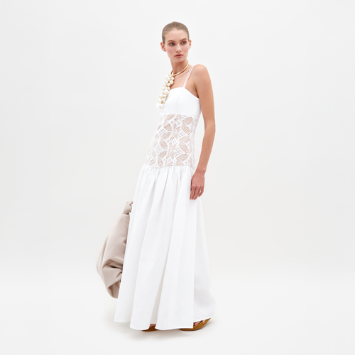 A woman stands wearing the Pilar Dress White, a sleeveless white dress featuring a lace midsection, and holds a beige bag against a plain white background. This elegant piece is available for pre-order and will be shipped by February 15th, 2025.