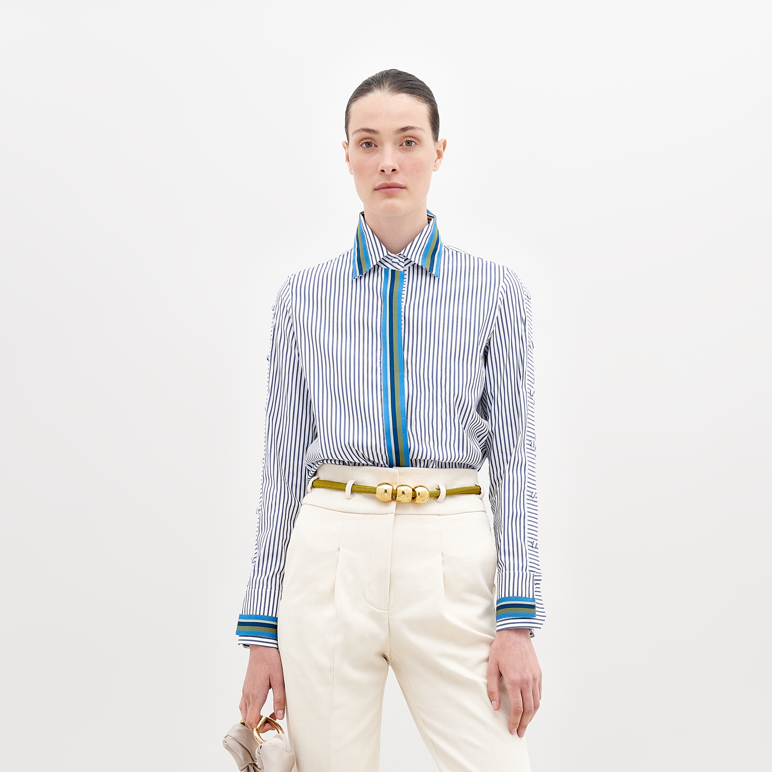 A person wearing a Premium Shirt White Blue Stripes with a blue accent, white pants, and a yellow belt holds a small bag in their hand. The background is plain white. This stylish ensemble will be available for pre-order starting November 15th, 2024.