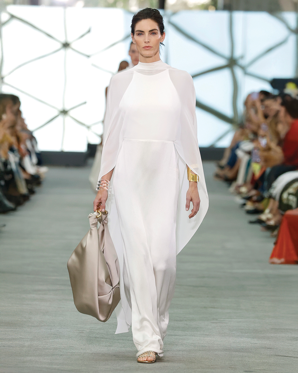Model walking on a fashion show runway wearing a white outfit.