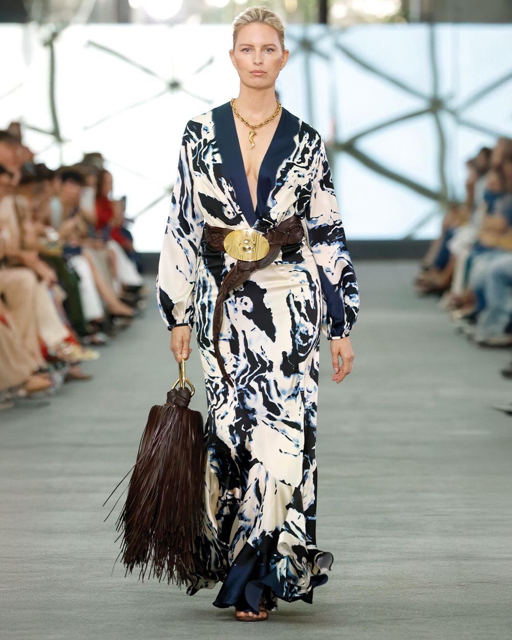 Model walking on a runway wearing a patterned dress with a belt and holding a broom.