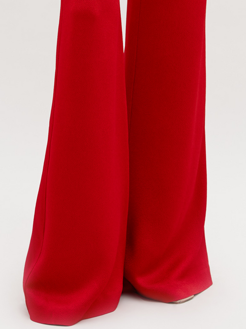 A person stands against a plain background wearing a red long-sleeved top with a knot detail at the waist and matching Palermo Pant Rouge, available for pre-order with a ship by date of November 15th, 2024.