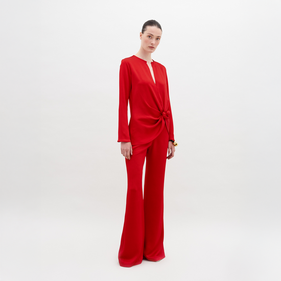 A person stands against a plain background wearing a red long-sleeved top with a knot detail at the waist and matching Palermo Pant Rouge, available for pre-order with a ship by date of November 15th, 2024.