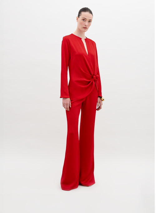 A person stands against a plain background wearing a red long-sleeved top with a knot detail at the waist and matching Palermo Pant Rouge, available for pre-order with a ship by date of November 15th, 2024.