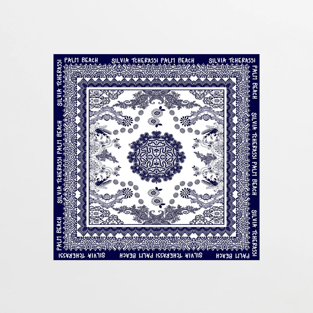 The Palm Beach Scarf is a square silk scarf featuring blue floral and geometric patterns on white, inspired by Mediterranean lace. Its blue border displays "PALM BEACH SILVIA TCHERASSI" in white lettering.