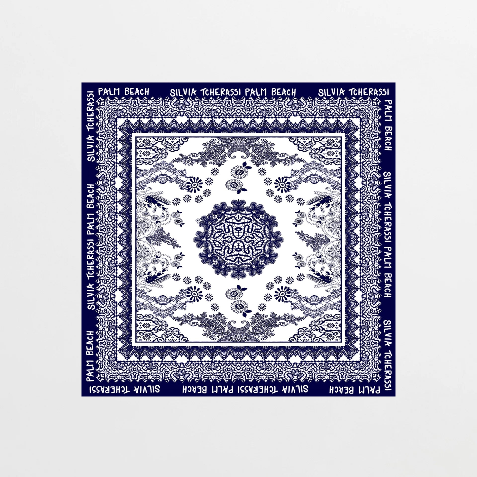 The Palm Beach Scarf is a square silk scarf featuring blue floral and geometric patterns on white, inspired by Mediterranean lace. Its blue border displays "PALM BEACH SILVIA TCHERASSI" in white lettering.