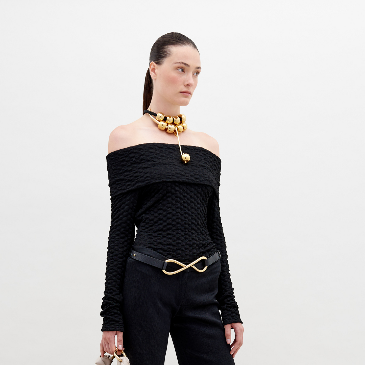 A woman is wearing an off-the-shoulder Palma Shirt Black, black pants with an infinity-shaped gold belt buckle, and a gold choker necklace with large round ornaments. She has her hair pulled back. Available for pre-order, this stylish ensemble will ship by November 15th, 2024.