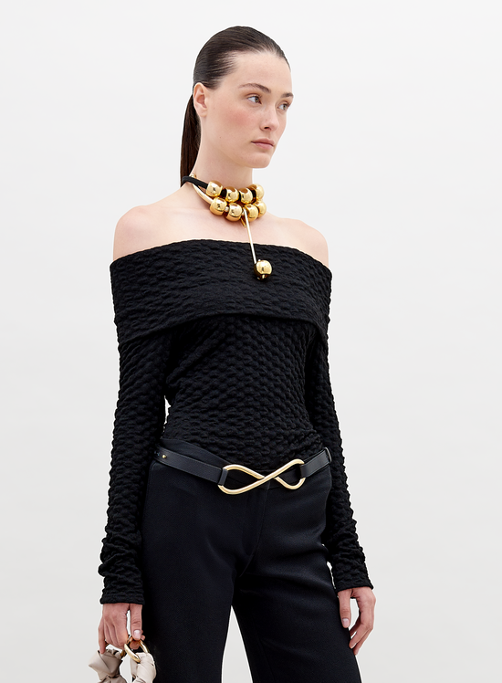 A woman is wearing an off-the-shoulder Palma Shirt Black, black pants with an infinity-shaped gold belt buckle, and a gold choker necklace with large round ornaments. She has her hair pulled back. Available for pre-order, this stylish ensemble will ship by November 15th, 2024.