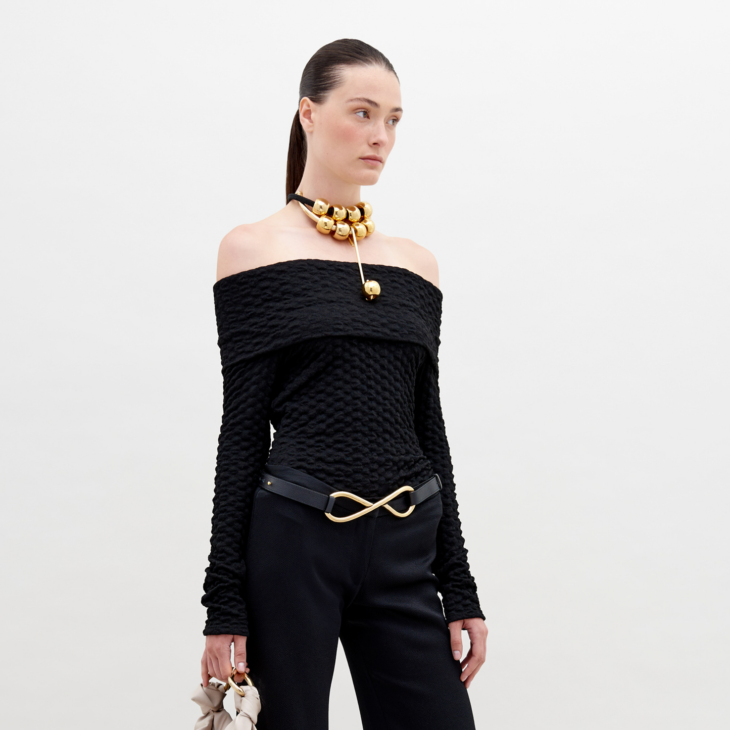 A woman is wearing an off-the-shoulder Palma Shirt Black, black pants with an infinity-shaped gold belt buckle, and a gold choker necklace with large round ornaments. She has her hair pulled back. Available for pre-order, this stylish ensemble will ship by November 15th, 2024.
