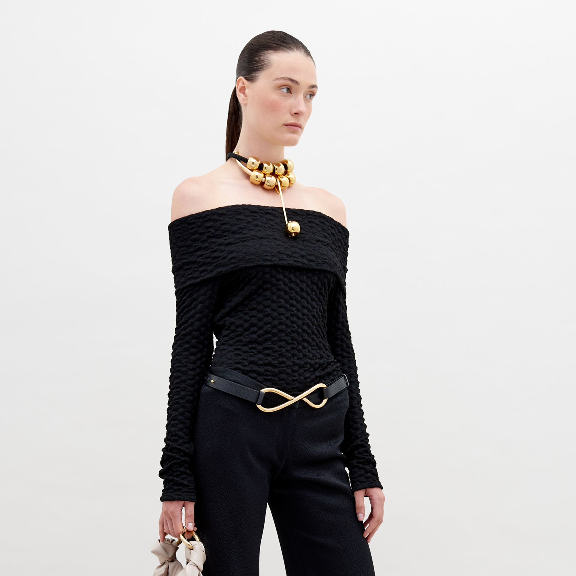A woman is wearing an off-the-shoulder Palma Shirt Black, black pants with an infinity-shaped gold belt buckle, and a gold choker necklace with large round ornaments. She has her hair pulled back. Available for pre-order, this stylish ensemble will ship by November 15th, 2024.