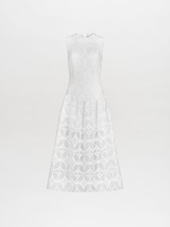 A person stands in the Paola Dress White, featuring a sleeveless lace design with a flared skirt, complemented by brown lace-up sandals. This item is available for pre-order and will ship by February 15th, 2025. The background is plain white.