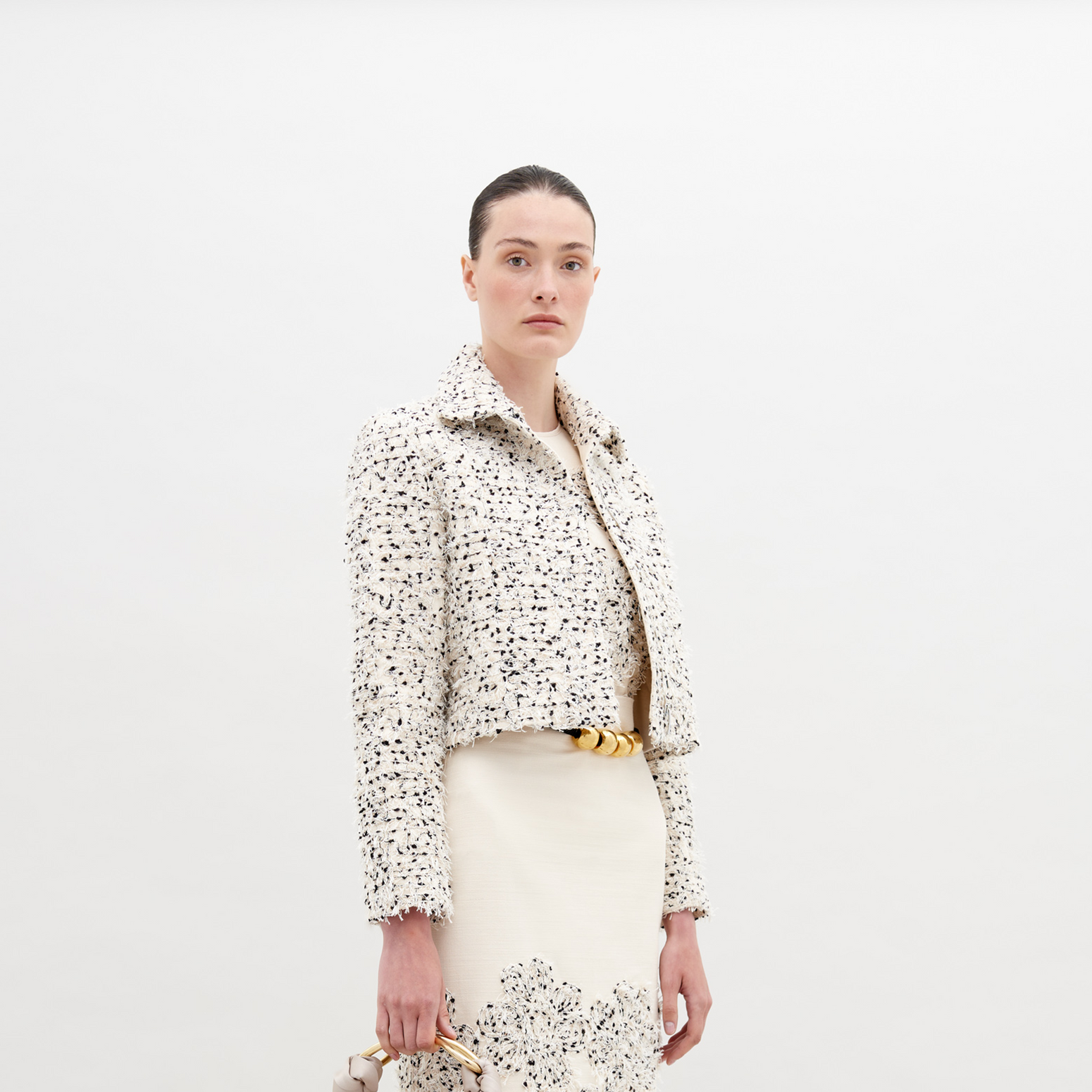 A woman with light skin and dark hair is wearing a Perla Jacket Beige Black Embroidered Flowers over a matching dress. Holding a small bag, she stands against a plain background. Pre-order now to ensure it ships by November 15th, 2024.