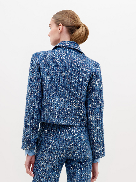 A person wearing a Perla Jacket Navy Sequins and pants, with a light blue shirt and a belt with pearl-like decorations, stands against a plain white background. Pre-order now to ensure delivery by November 15th, 2024.