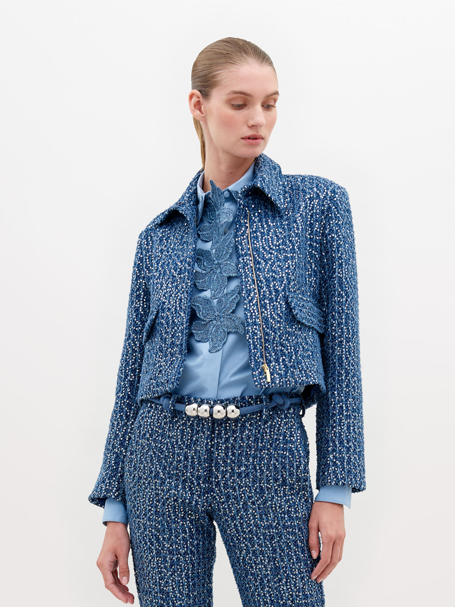 A person wearing a Perla Jacket Navy Sequins and pants, with a light blue shirt and a belt with pearl-like decorations, stands against a plain white background. Pre-order now to ensure delivery by November 15th, 2024.