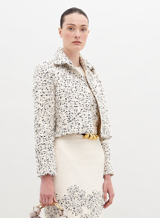 A woman with light skin and dark hair is wearing a Perla Jacket Beige Black Embroidered Flowers over a matching dress. Holding a small bag, she stands against a plain background. Pre-order now to ensure it ships by November 15th, 2024.