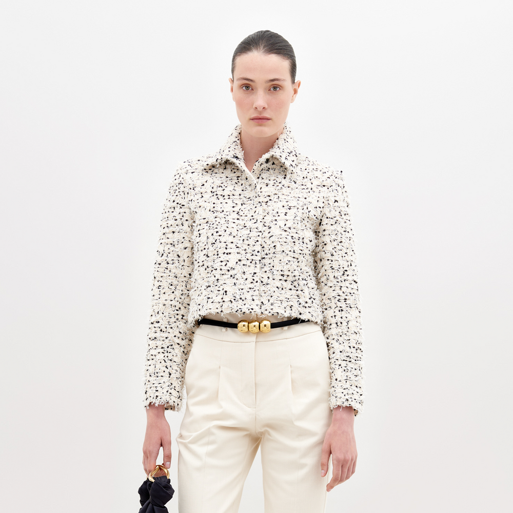 A woman with light skin and dark hair is wearing a Perla Jacket Beige Black Embroidered Flowers over a matching dress. Holding a small bag, she stands against a plain background. Pre-order now to ensure it ships by November 15th, 2024.