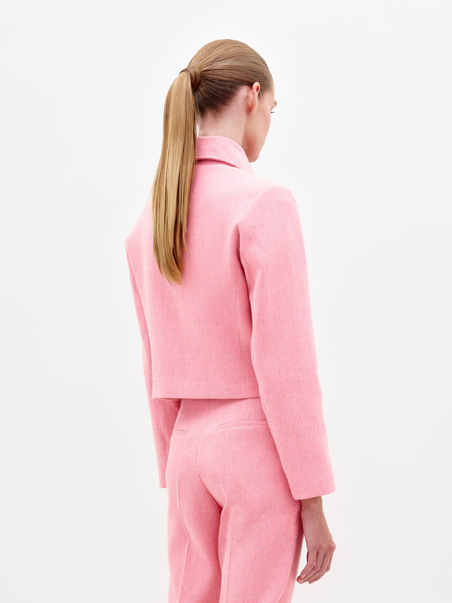 A person with tied-back hair is wearing a matching pink tailored suit, including the Perla Jacket Pink, which features a collar and two front pockets, standing against a plain white background. This stylish look will be available for pre-order, with the ship date set for February 2025.