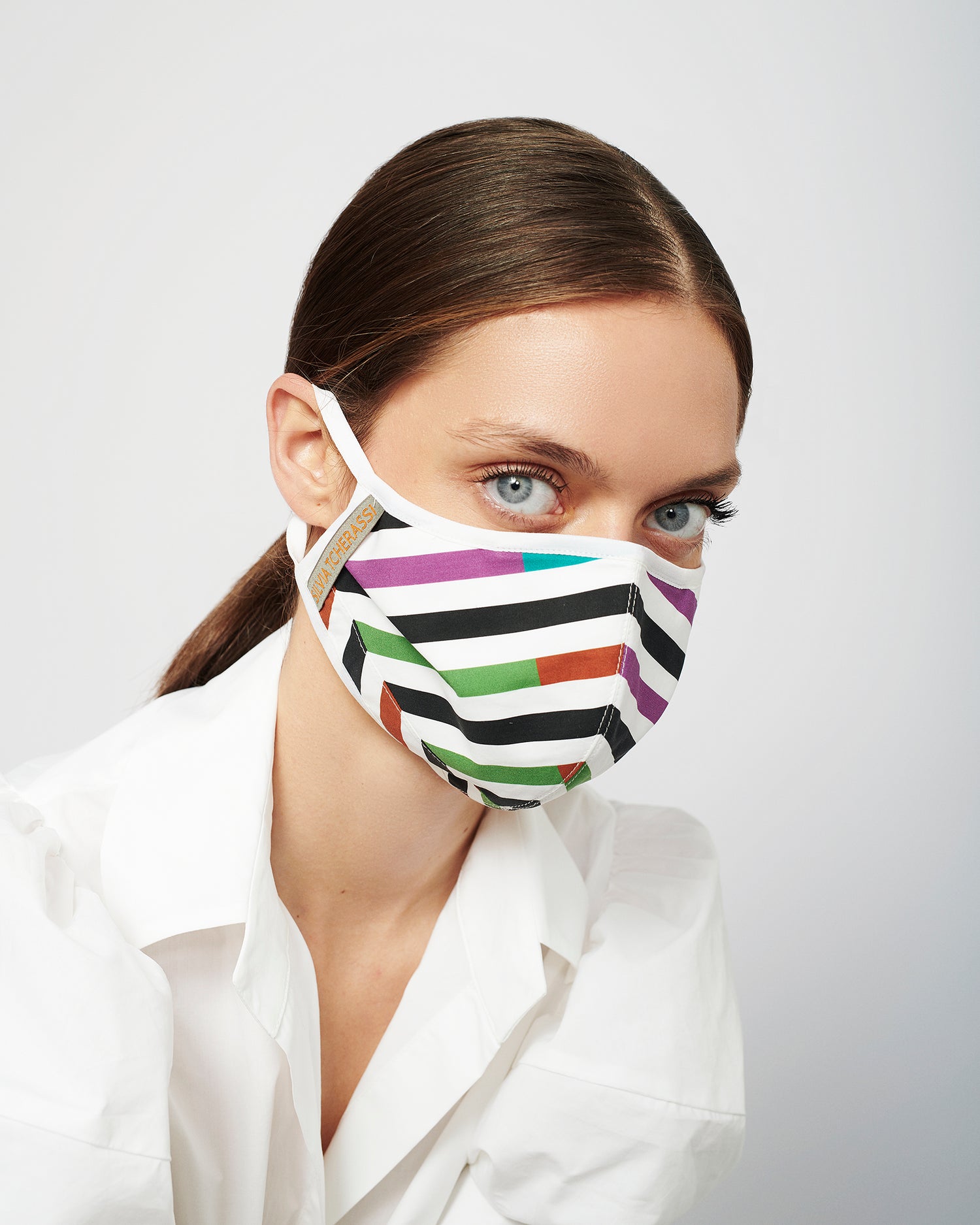 Person wearing a striped, colorful face mask and a white shirt, looking directly at the camera against a plain background.