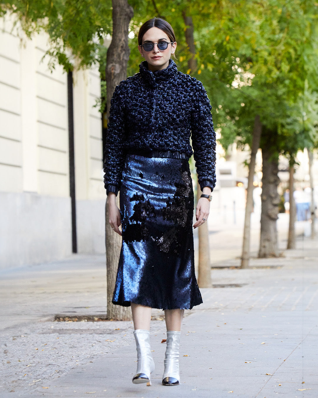 Person wearing a textured black top, shiny black skirt, silver boots, and sunglasses, standing on a sidewalk lined with trees.