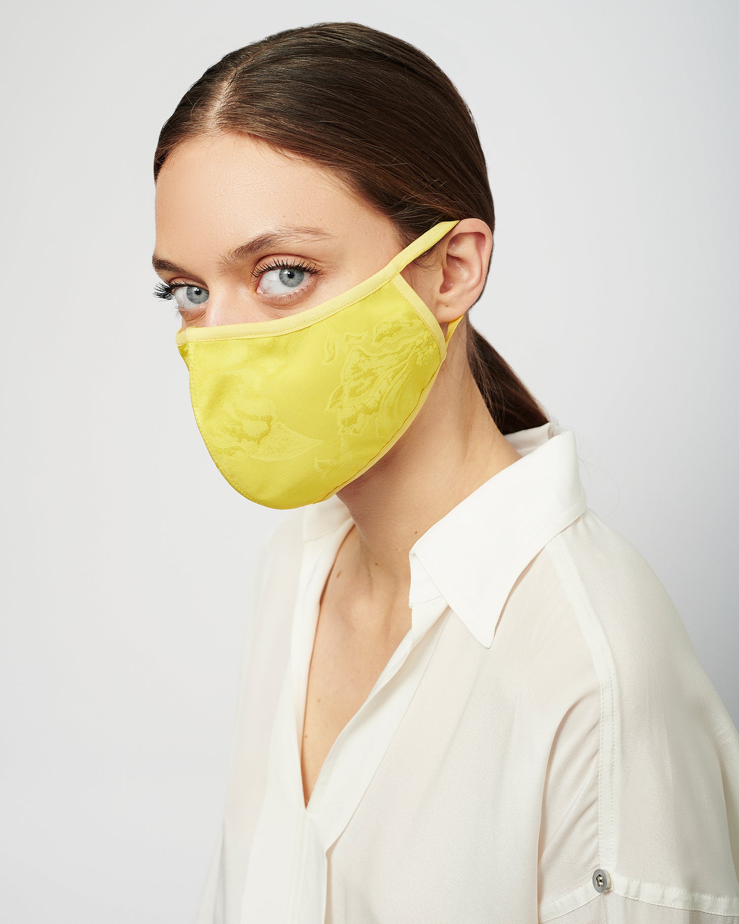 Person wearing a yellow face mask and a white shirt looks towards the camera against a plain background.