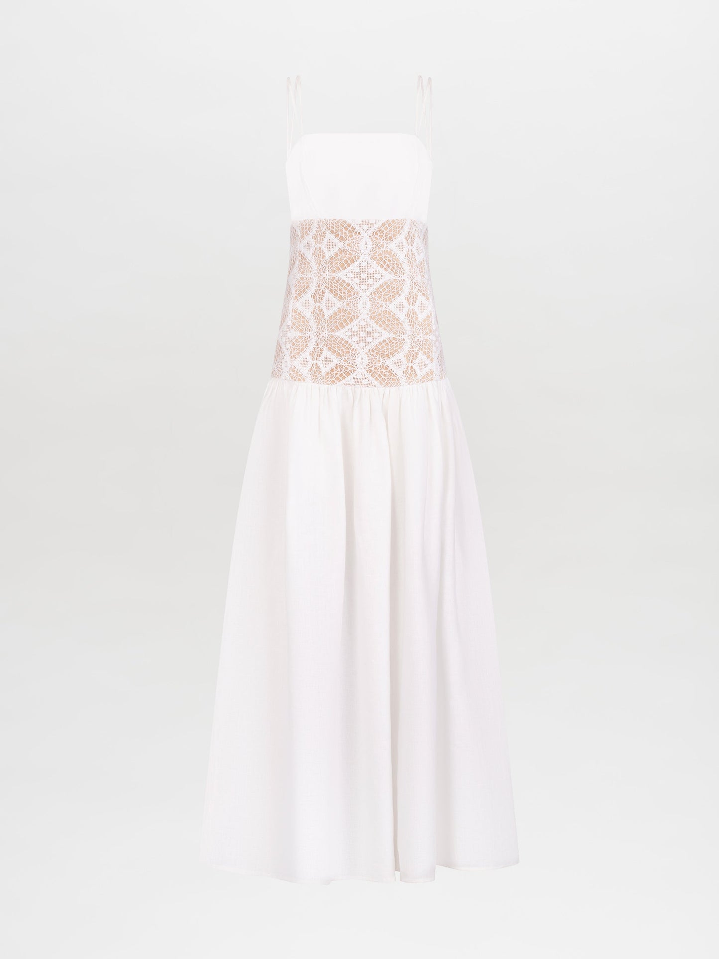 A woman stands wearing the Pilar Dress White, a sleeveless white dress featuring a lace midsection, and holds a beige bag against a plain white background. This elegant piece is available for pre-order and will be shipped by February 15th, 2025.