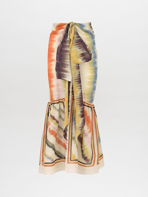 The Portia Skirt in Multi Hazy Stripe design features a long silhouette crafted from lightweight cotton voile, accented by a self-tie waistband and adorned with a tie-dye pattern in vibrant shades of orange, yellow, and blue on a light background.