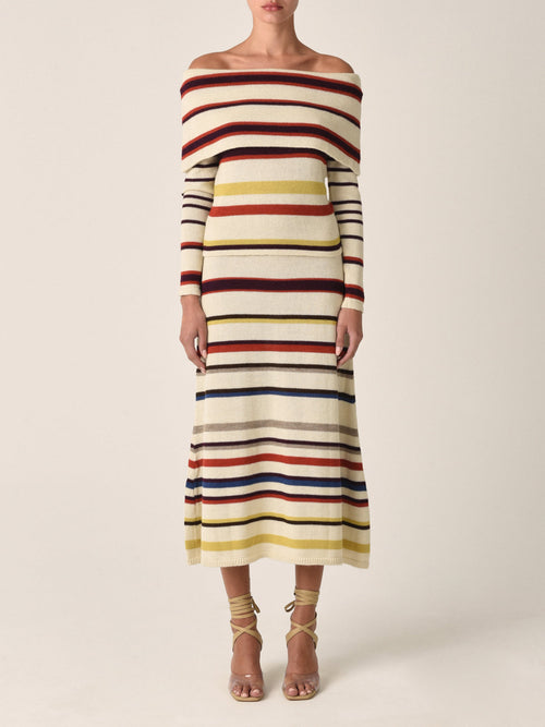 A person models the Brie Skirt Multi Mustard Stripe, featuring an off-the-shoulder design with long sleeves and ankle-length cut. Crafted from 100% baby alpaca and sustainably dyed, this striped knit piece is a standout choice that pairs perfectly with strappy sandals.