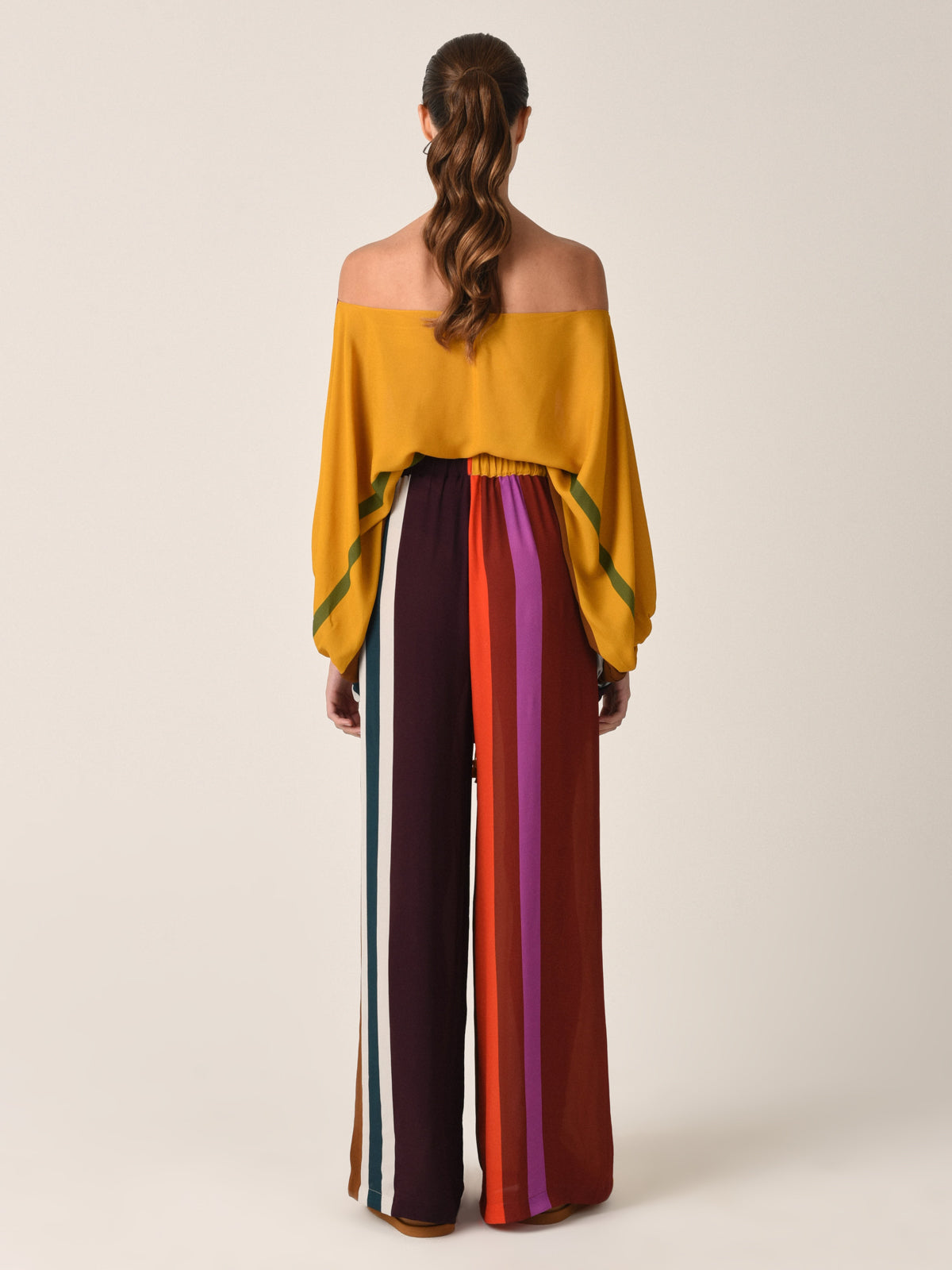 The Bellagio Blouse Color Block Multi is a chic, off-shoulder top with draped sleeves, adorned with horizontal stripes in vibrant shades of red, purple, orange, and brown. Its multi-colored design and flattering silhouette make it a stylish choice for any occasion.