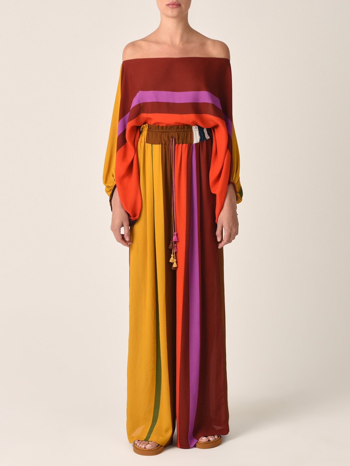 The Ovada Pant Color Block Multi features wide-leg high-waisted trousers with vertical stripes in yellow, orange, red, purple, pink, and green. These pants include an elastic waistband with a drawstring tie and are crafted from soft viscose fabric.